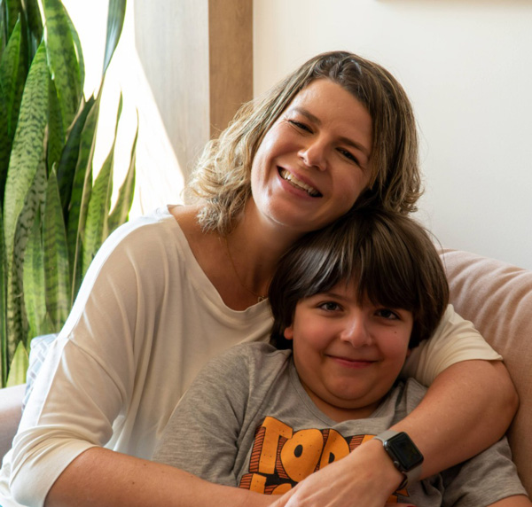 Foto de Alessandra, que é uma mulher de cabelos enrolados e claros na altura dos ombros, usando uma camiseta branca, sentada no sofá e sorrindo abraçada a seu filho, um garoto de uns 11 anos, com cabelos castanhos, usando camiseta cinza e também sorrindo.