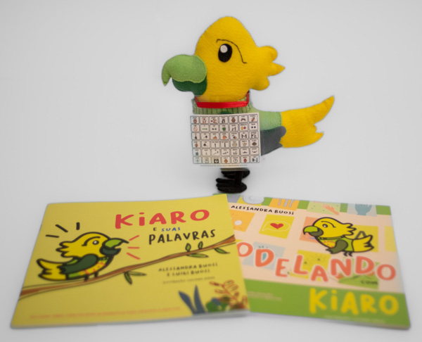 Foto do papagaio Kiaro feito em feltro, em um fundo branco com dois livros em sua frente: "Kiaro e suas palavras" e "Modelando com Kiaro"