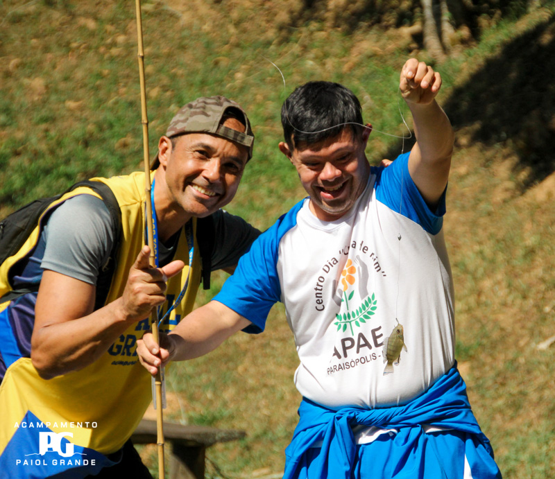 Garoto com camiseta da APAE segundando uma vara de pesca e uma linha com um peixinho na ponta. Ao seu lado um monitor do acampamento com a camiseta do Paio Grande.