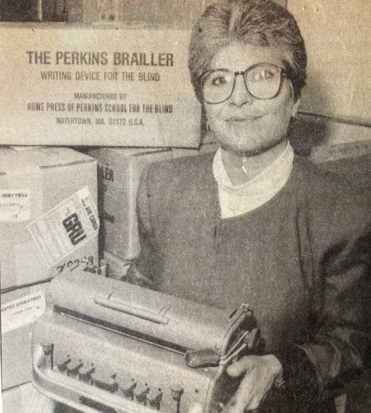 Foto de jornal antiga, com uma senhora de óculos, cabelos claros e curtos, segurando uma máquina de escrever braille. Atrás dela diversas caixas.