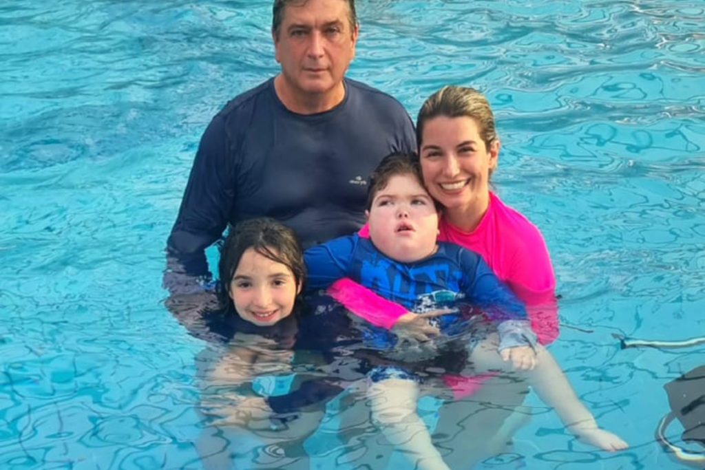 Foto de um pai, uma mãe, uma garotinha de 10 anos e um garotinho de 6 anos no colo da mãe. Todos estão dentro de uma piscina bem azul.