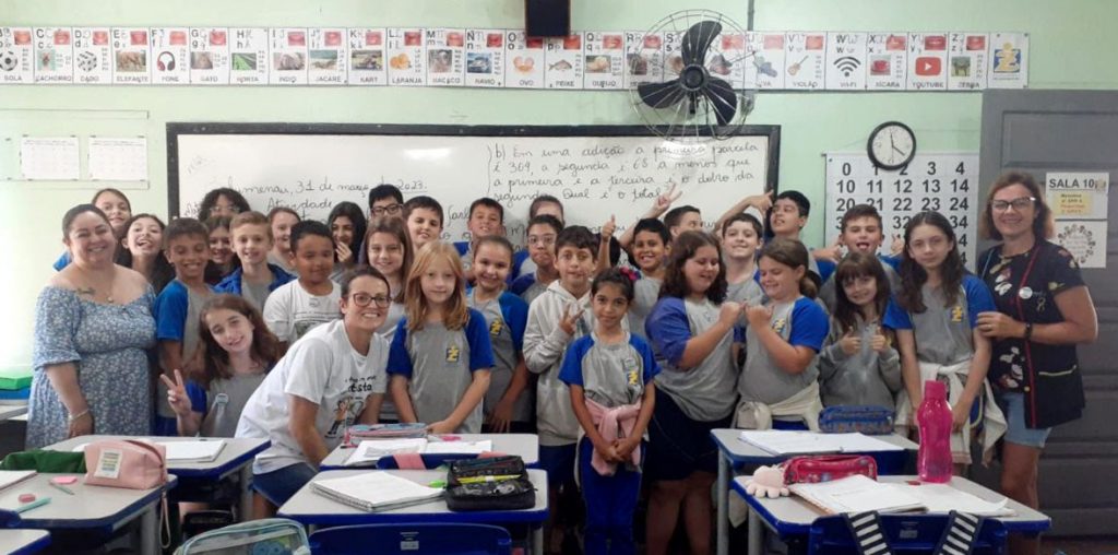 Foto de uma sala de aula, com todos os alunos da classe posando para a foto em frente a lousa. As professoras estão ao lado.