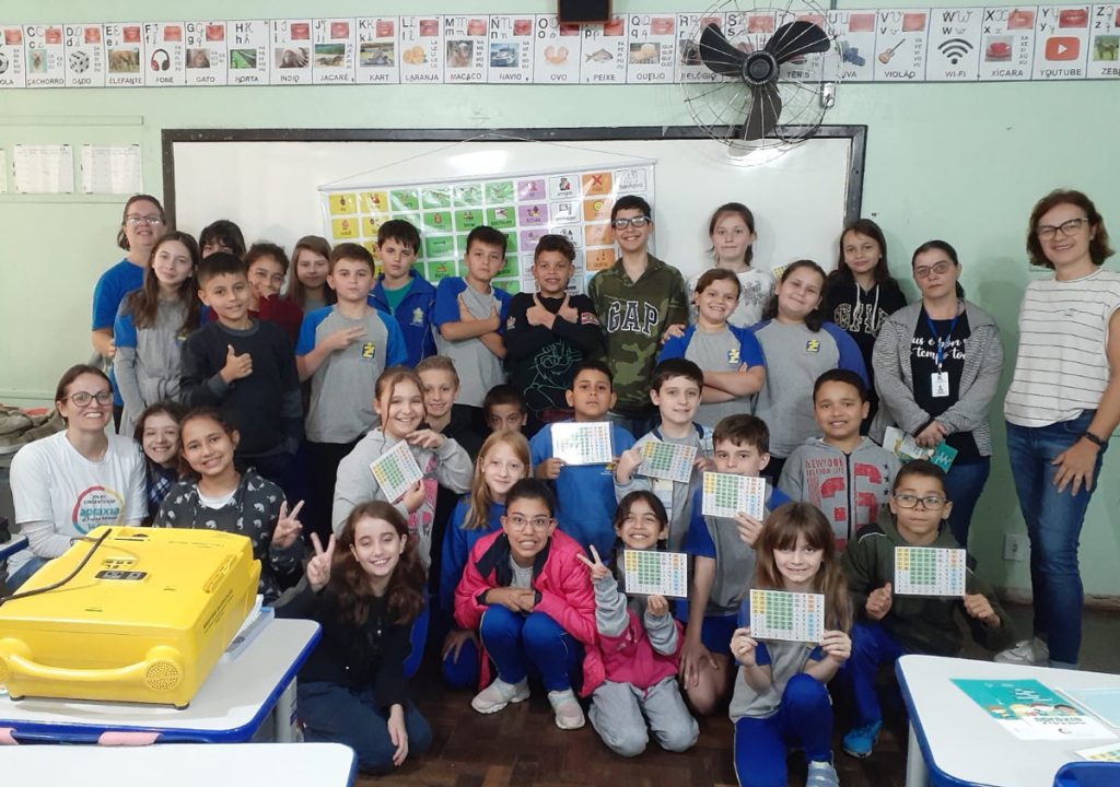 Foto da classe do Pedro com vários alunos segurando uma prancha de comunicação alternativa. Na foto também estão as duas professoras e Giselle, mãe do Pedro.