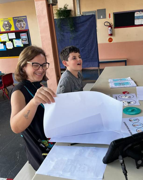 Foto de uma escola, com um aluno (Pedro) sentado em uma mesa fazendo atividades com sua professora (Lenira) ao lado.