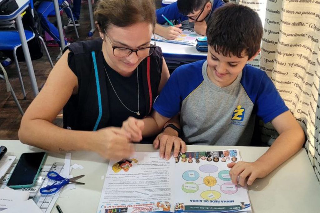 Foto de uma sala de aula. Em primeiro plano uma carteira com um aluno (Pedro) sentado com sua professora (Lenira), fazendo uma atividade. Ao fundo outras carteiras e alunos.