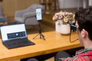 Foto de uma mesa com um notebook e um celular, em frente a mesa vemos de costas um rapaz usando um óculos com um dispositivo fixo na lateral e um acionador de mordida.