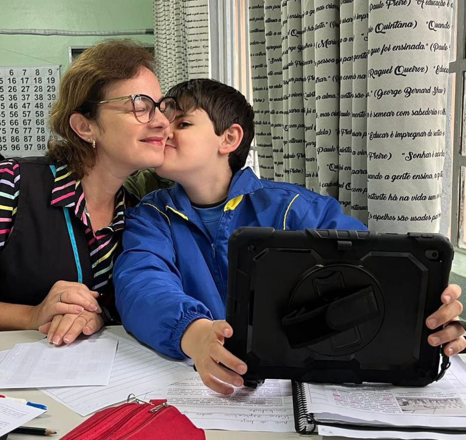 Foto de uma sala de aula, onde o aluno Pedro está sentado em uma carteira, segurando um tablet e dando um beijo em sua professora que está sentada ao seu lado.