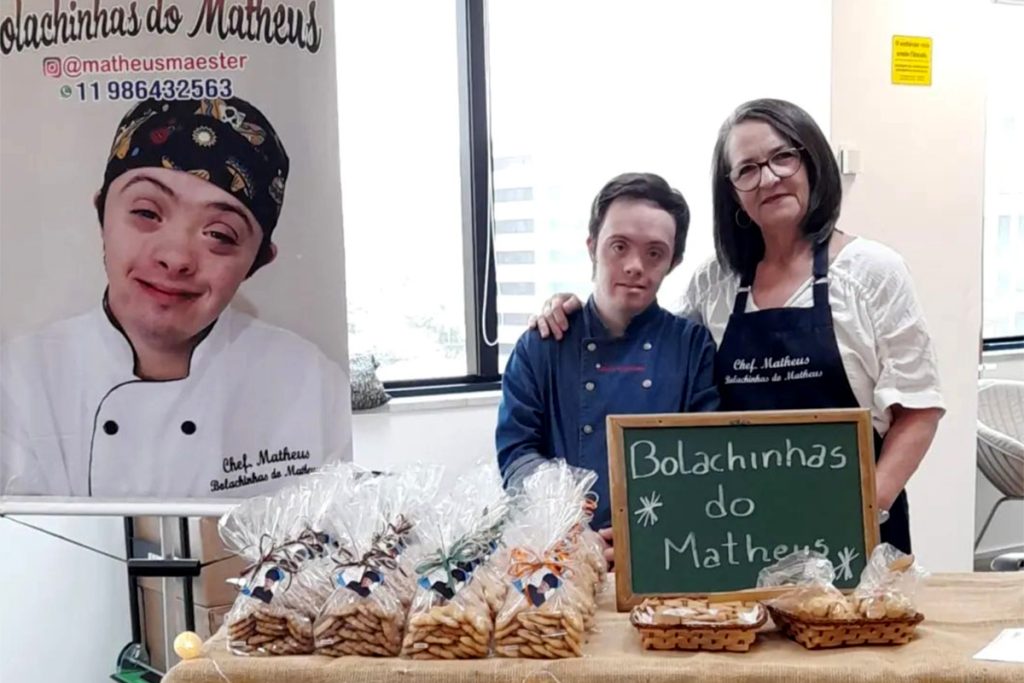 Foto de Matheus e sua mão com roupas de "chef" atrás de uma mesa com várias embalagens de bolachinhas e uma lousa escrito: Bolachinhas do Matheus.