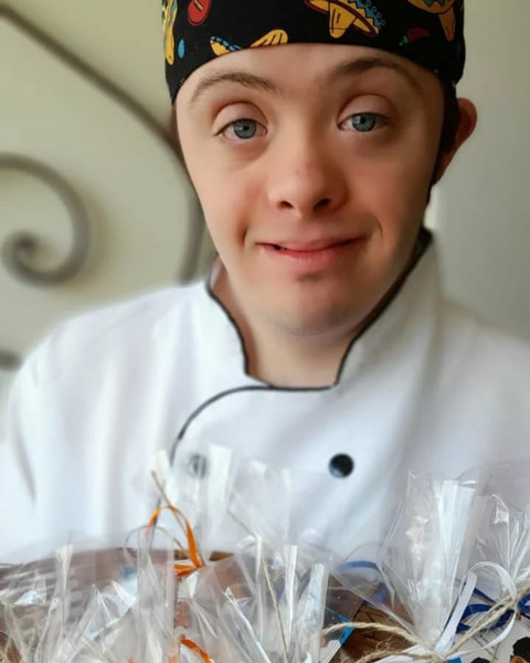 Foto em close de Matheus, que é um garoto com olhos claros e sorridente, usando doma e bandana de "chef", segurando uma bandeja cheia de embalagens com bolachinhas.