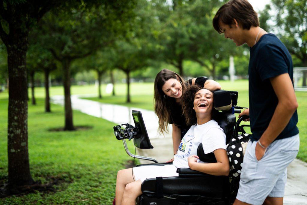 Foto de uma garota jovem, morena, em uma cadeira de rodas, com seu tablet de comunicação assistiva em sua frente, está em um parque com mais dois amigos. Todos estão se divertindo.