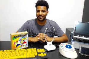 Foto de Carlos, um rapaz jovem, moreno, com barba, usando uma camiseta cinza, sentado em uma mesa preta com diversos recursos de tecnologia assistiva em sua frente.