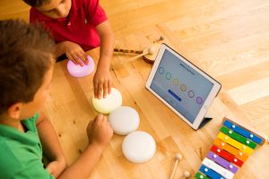 Foto vista de cima de dois meninos interagindo com o Cosmo (4 botões brancos que acendem luzes), seguindo a orientação do tablet que está na frente deles. Sobre a mesa também tem um xilofone colorido e um tamborzinho.