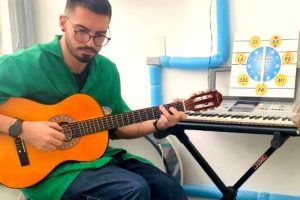 Foto do musicoterapeuta Pedro Della Rosa na sala de atendimento, tocando violão, e com um teclado ao lado.