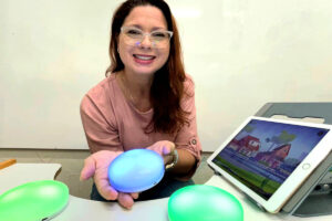 Foto de Tathiane Cuesta sorrindo apoiada em uma mesa e segurando em suas mãos um "cosmóide", na mesa vemos um tablet com uma imagem gráfica de cidade e outros dois cosmóides