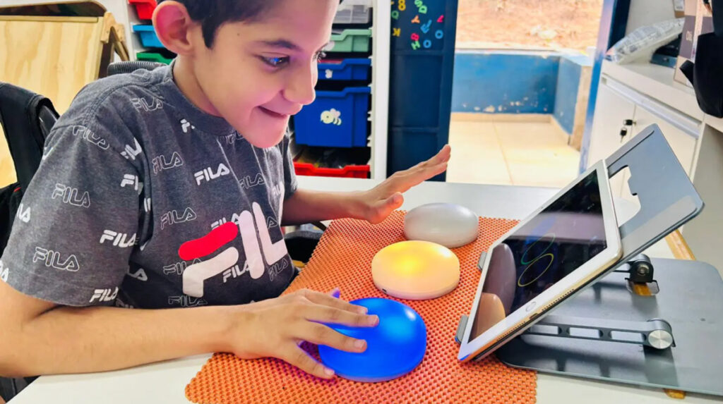 Garoto sorrindo, sentado em uma carteira escolar. Em sua frente tem um tablet e 3 cosmóides (botões grandes coloridos). Ele está apertando o botão azul.