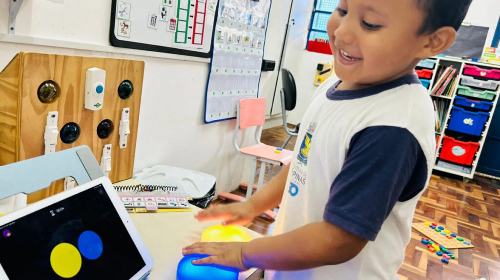 Foto de um garotinho moreno sorrindo em uma sala de aula. Em sua frente tem uma mesa com dois cosmóides (botões grandes e coloridos) e um tablet. Ele está apertando o botão azul.