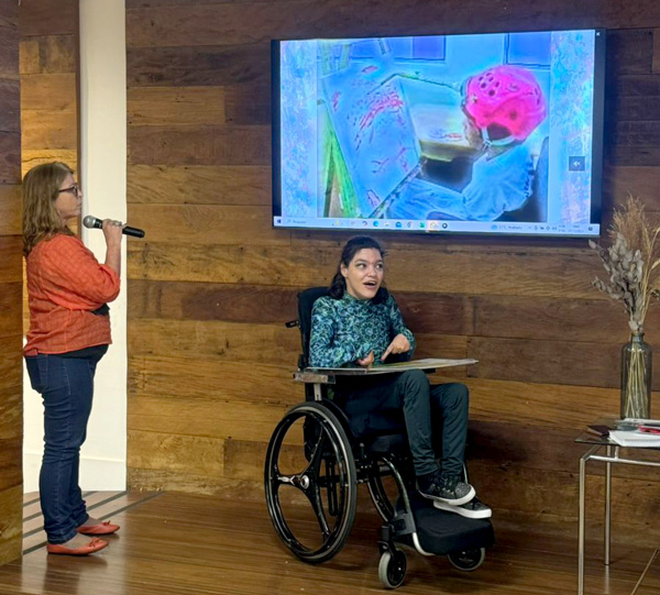 Foto de Samara no palco em sua cadeira de rodas junto com sua mãe Claudia com microfone na mão e no alto uma TV com uma tela da apresentação com a foto de Samara mais nova pintando com uma ponteira de cabeça