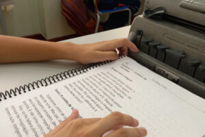 Foto em close de uma mesa de escola com uma apostila com um texto normal e em braille. a mão do aluno está passando sobre as letras braille. Sobre a mesa uma máquina de escrever braille