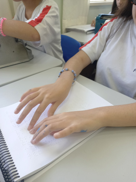 Foto em close de uma sala de aula onde vemos em primeiro plano uma aluna lendo com os dedos um caderno em braille