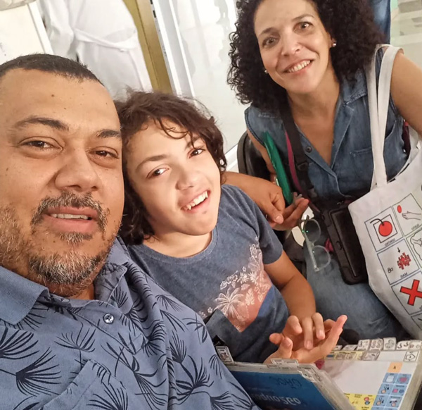 Foto de Shirley, Rui e João olhando e sorrindo para a foto. João está com um livro de CAA em seu colo.