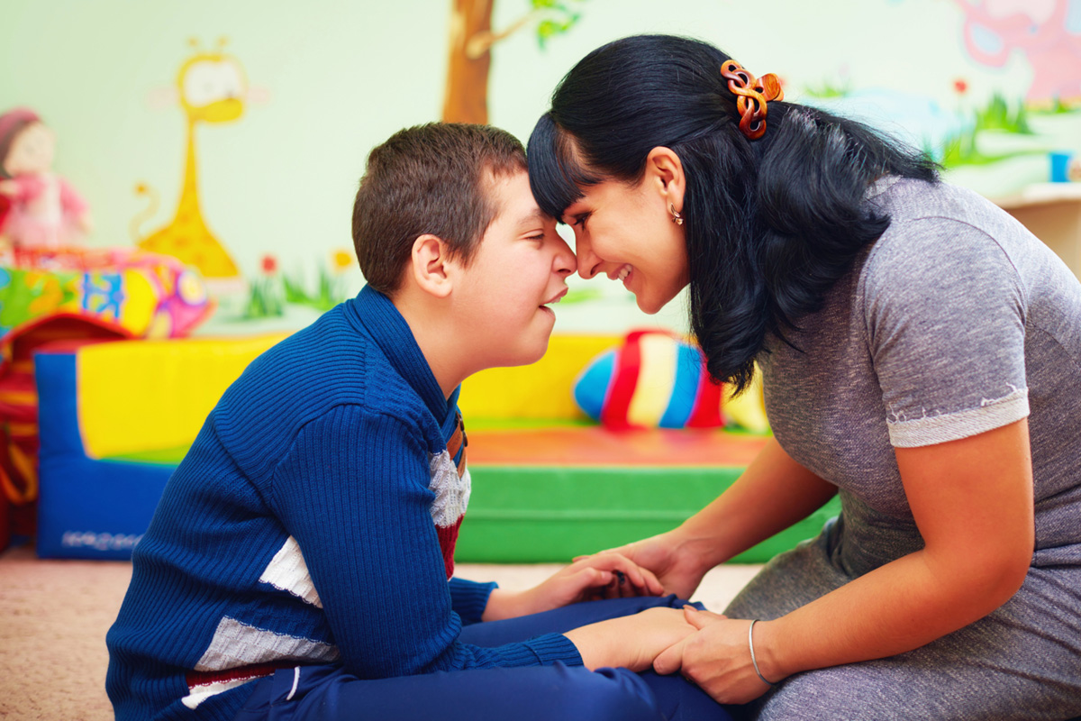 Foto de uma mãe sentada no chão sorrindo e segurando as mãos de seu filho com deficiência. Ao fundo vemos desfocado uma sala com decoração infantil