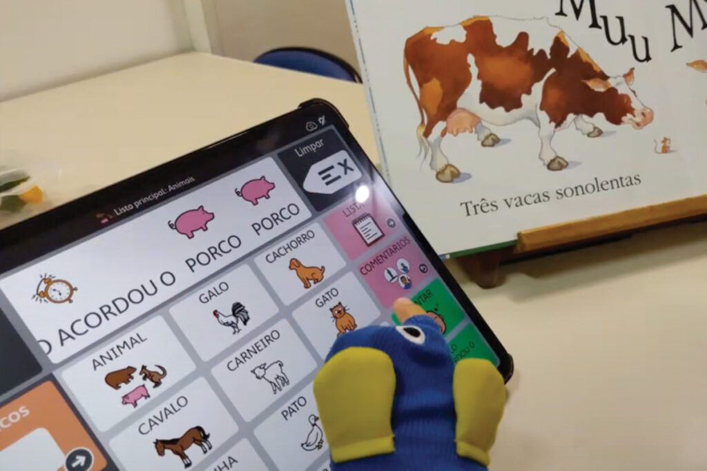 Foto de uma mesa, com um livro chamado 3 vacas sonolentas, e ao lado um tablet de Comunicação Alternativa está com o texto do livro em pictogramas