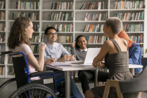 Foto de uma biblioteca com uma mesa retangular com livros e cadernos sobre ela e em volta 5 estudantes universitários, sendo uma jovem cadeirante.