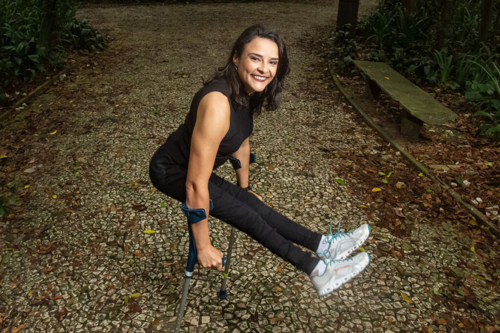 Foto de Jessica em um parque sorrindo e se equilibrando em suas muletas.