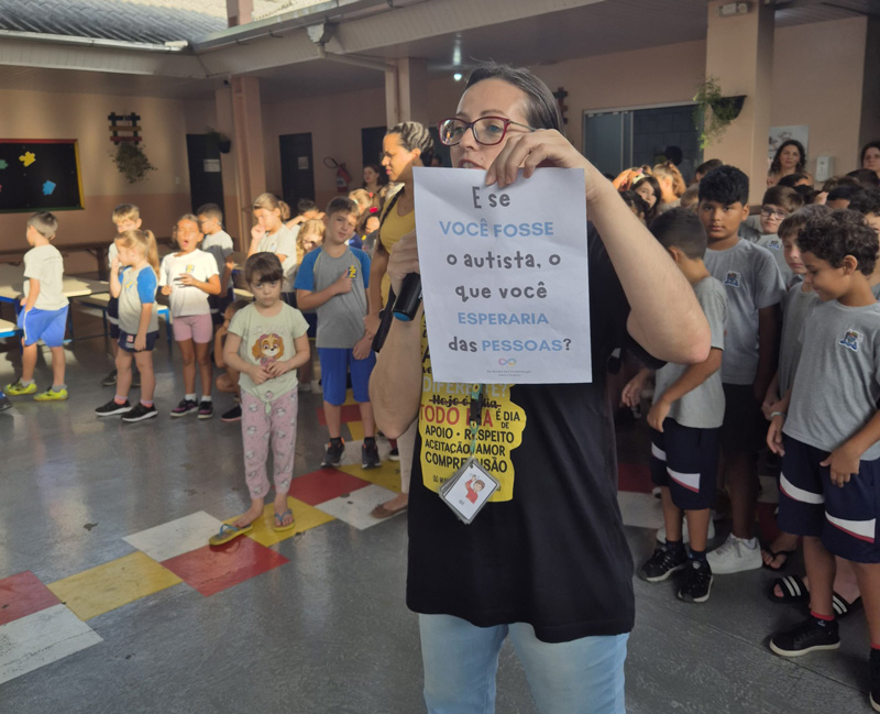 Foto de Giselle no pátio da escola do seu filho com uma folha escrito: E se você fosse o autista, o que você esperaria das pessoas?