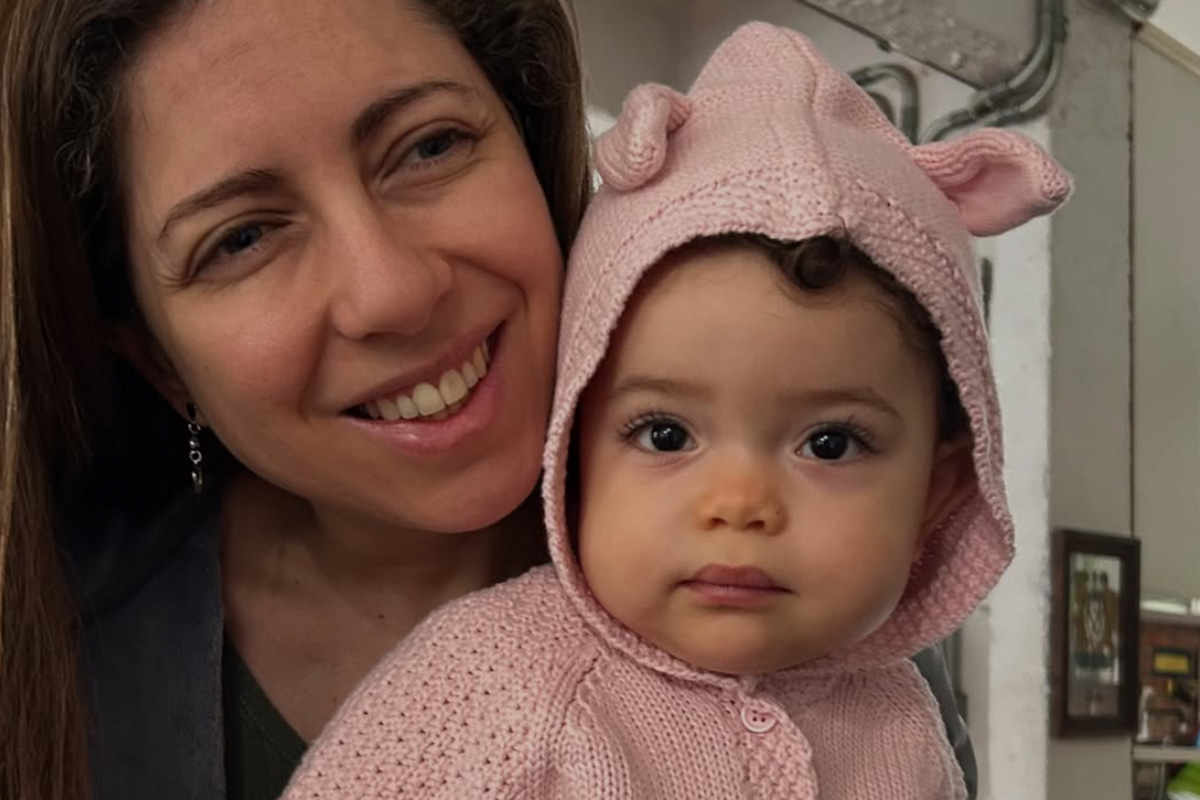 Foto de Mel sorrindo com sua filha Sofia no colo vestindo um casaquinho rosa com capuz.