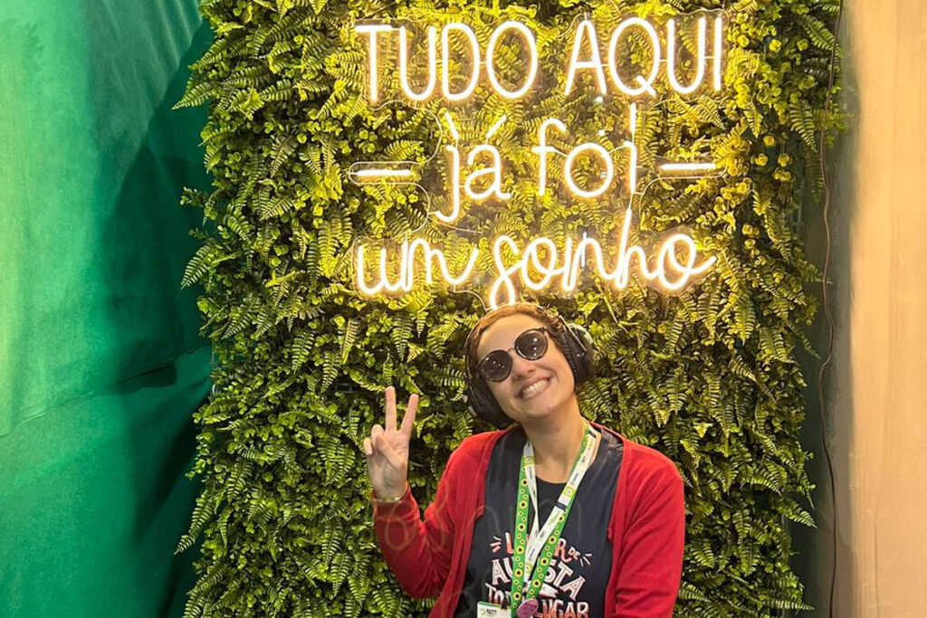 Foto de Fernanda sorrindo e fazendo sinal de "paz e amor" com as mãos sentada em frente a um luminoso escrito: "Tudo aqui já foi um sonho"