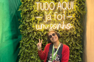 Foto de Fernanda sorrindo e fazendo sinal de "paz e amor" com as mãos sentada em frente a um luminoso escrito: "Tudo aqui já foi um sonho"