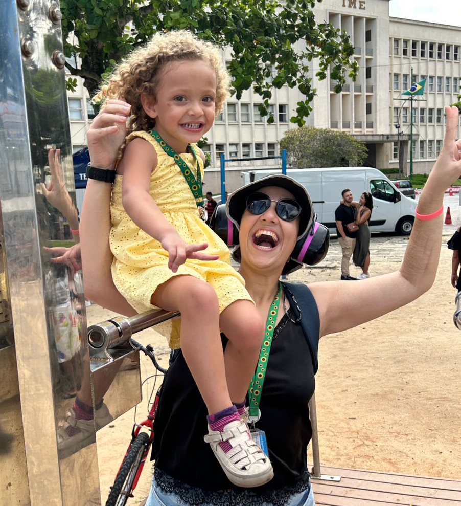 Foto de Fernanda e sua filha Ana Laura em um aparelho de ginástica em uma praia do Rio sorrindo para a câmera. Ambas usam o cordão de girassol.