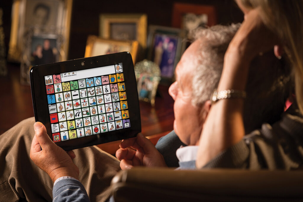 Foto de um senhor segurando sentado em uma sala segurando um tablet de CAA. Sua esposa está atrás dele, olhando o que ele está querendo falar.