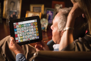 Foto de um senhor segurando sentado em uma sala segurando um tablet de CAA. Sua esposa está atrás dele, olhando o que ele está querendo falar.