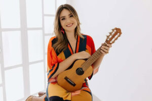 Foto de Elisa Gatti sentada no chão sorrindo segurando um pequeno violão
