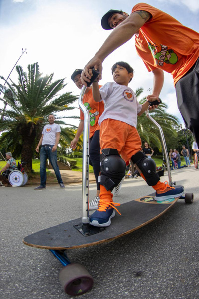 Foto de um garotinho andando em um skate adaptado com a ajuda de dois profissionais