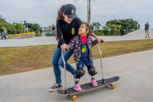 Foto de uma garotinha se divertindo ao andar em um skate adaptado com a ajuda de uma profissional