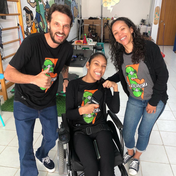 Foto de Nayumi em sua cadeira de rodas com a camiseta do SkateAnima em uma sala de fisioterapia ao lado de Stevan e sua mãe