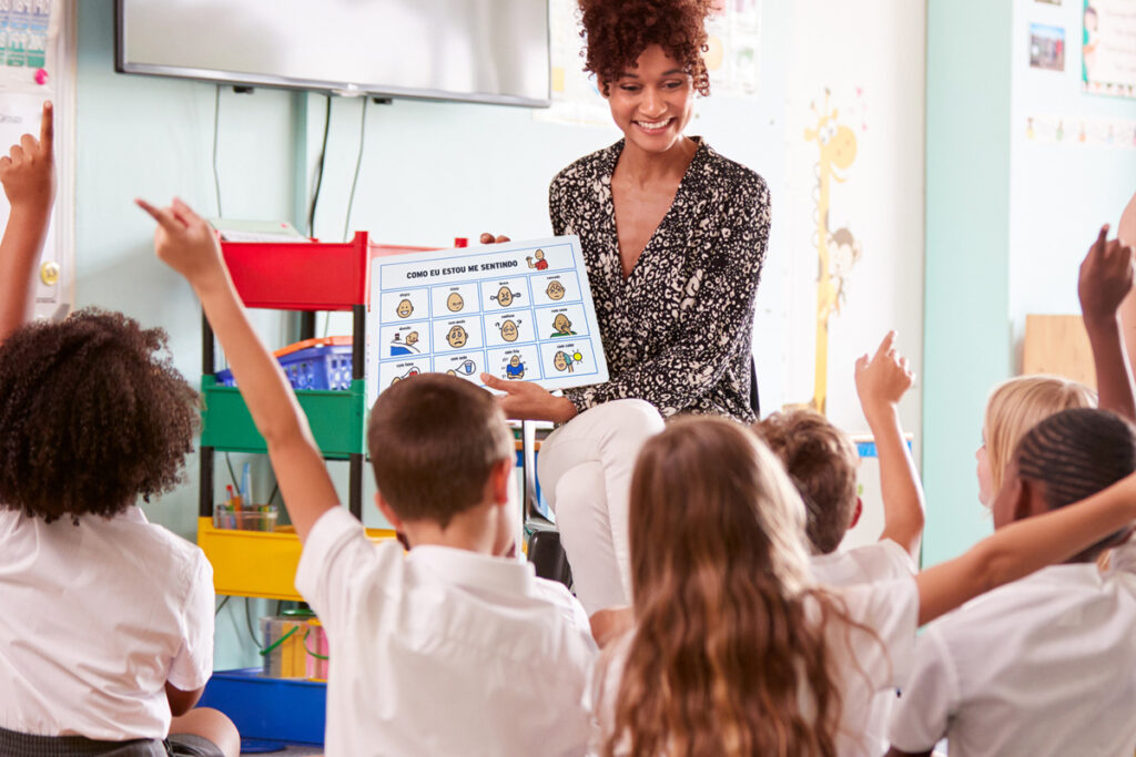 Foto de uma professora mostrando uma prancha de Comunicação Alternativa sobre sentimentos para seus alunos que estão sentados em no chão em sua frente. Alguns estão com a mão levantada para querer falar.