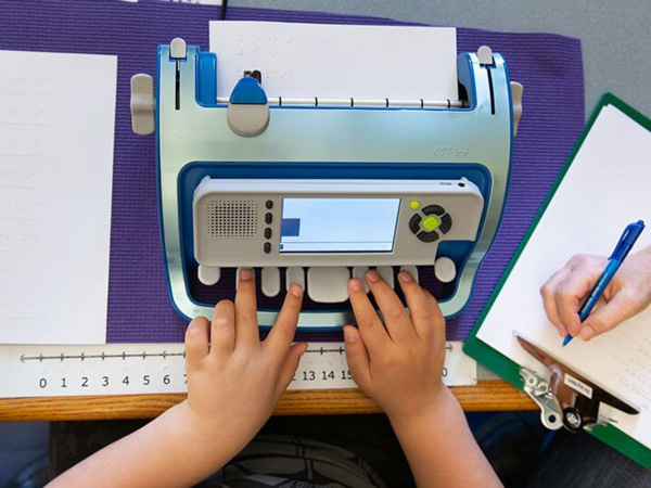 Foto em close de uma máquina de escrever braille com uma criança apertando suas teclas. Ao lado uma prancheta com outra pessoa escrevendo.