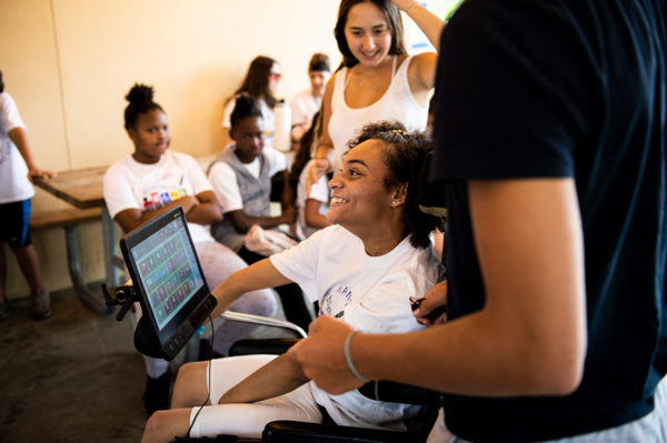 Foto de uma jovem em uma cadeira de rodas com seu tablet com rastreador ocular em sua frente. Ela está sorrindo em um ambiente escolar.