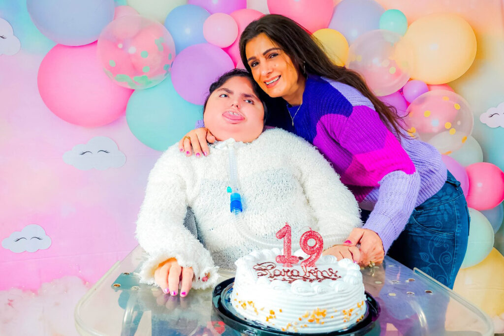 Foto da Helo sentada em frente a um bolo com vela de 19 anos. Abraçada a ela está sua mãe.