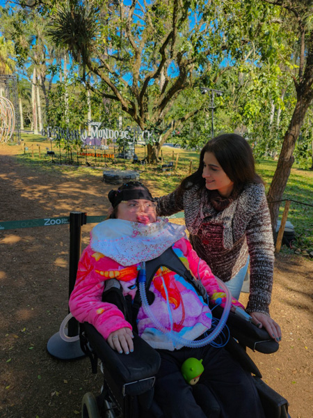 Foto de Helo em sua cadeira de rodas junto com sua mãe no zoologico.