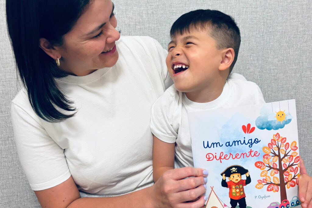 Foto de Priscila sorrindo, olhando para seu filho Ben em seu colo. Ela está segurando o livro que escreveu: "Um amigo diferente"