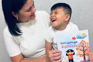 Foto de Priscila sorrindo, olhando para seu filho Ben em seu colo. Ela está segurando o livro que escreveu: "Um amigo diferente"