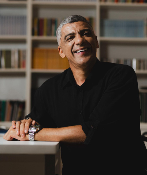 Foto de Geraldo sorrindo com camisa preta e ao fundo uma estante de livros.
