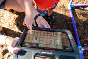 Foto de uma criança com a mãe em um parque, e está em close um tablet de Comunicação Alternativa qe a mãe está segurando, onde a criança aponta uma figura