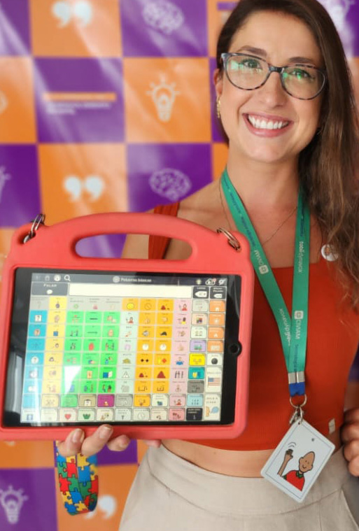 Foto da fonoaudiologa Natalia sorrindo, segurando um tablet de Comunicação Alternativa e usando um colar de CAA