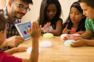 Foto de uma turma de alunos ao redor de uma mesa jogando um jogo interativo junto com o professor